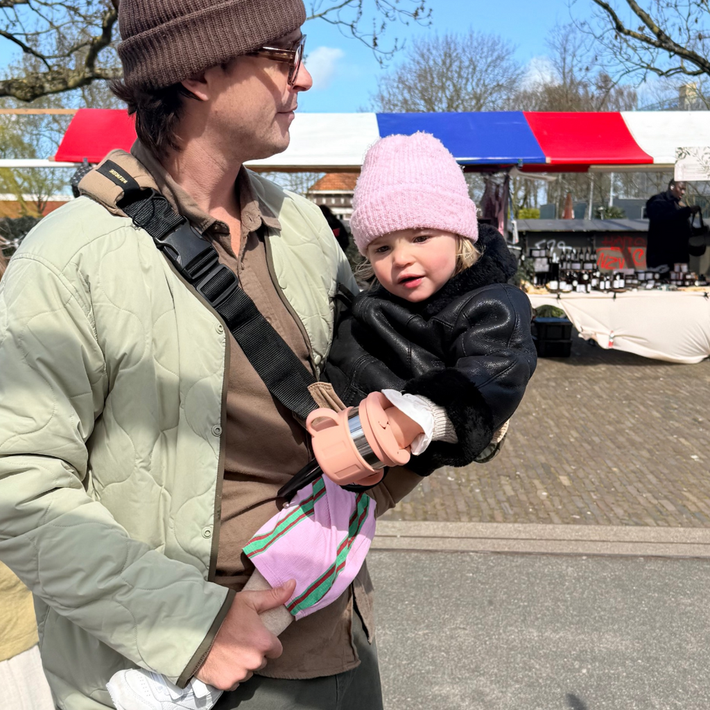 Man holding a child in a toddler carrier in an outdoor setting with a colorful canopy in the background and a mounted pink snack cup