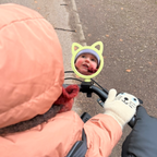 Child in a pink coat looking into a green mirror with cat ears on a bicycle.