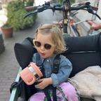 Child in a bakfiets eating a snack from a pink mounted snack cup, wearing sunglasses with bicycles in the background.